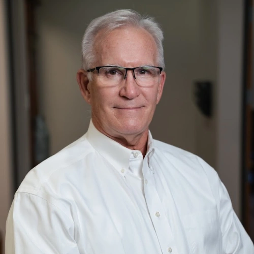 Professional headshot of older man with white hair and glasses wearing white button-up shirt against neutral background.